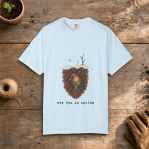 See You in Spring gardening t-shirt in Chambray, laid flat on a potting bench with gardening gloves and a trowel
