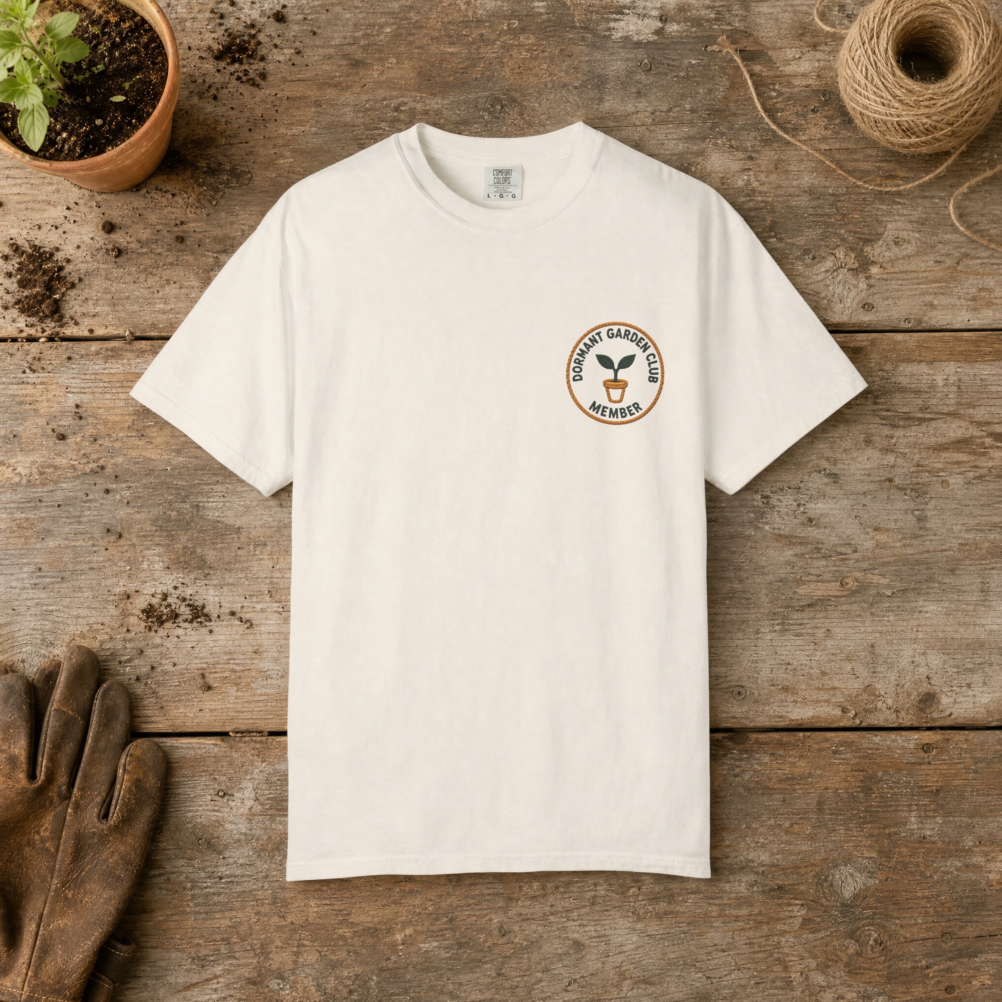 Dormant Garden Club Member gardening t-shirt in Ivory, laid flat on a potting bench with gardening gloves and a trowel
