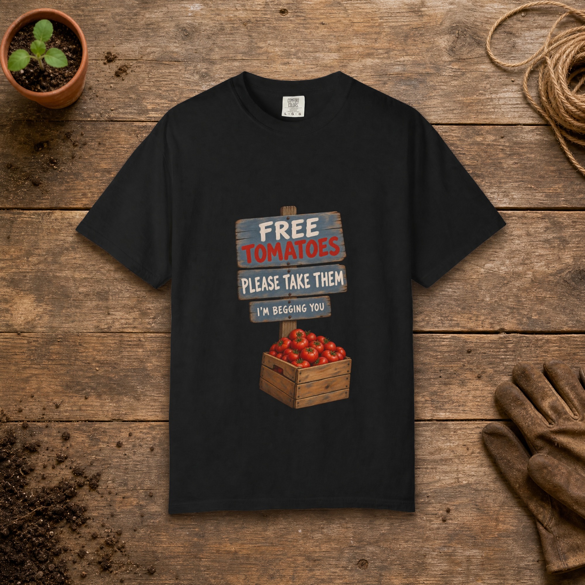 Free Tomatoes gardening t-shirt in Black, laid flat on a potting bench with gardening gloves and a trowel