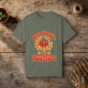 Bad Knees, Great Tomatoes gardening t-shirt in Moss, laid flat on a potting bench with gardening gloves and a trowel