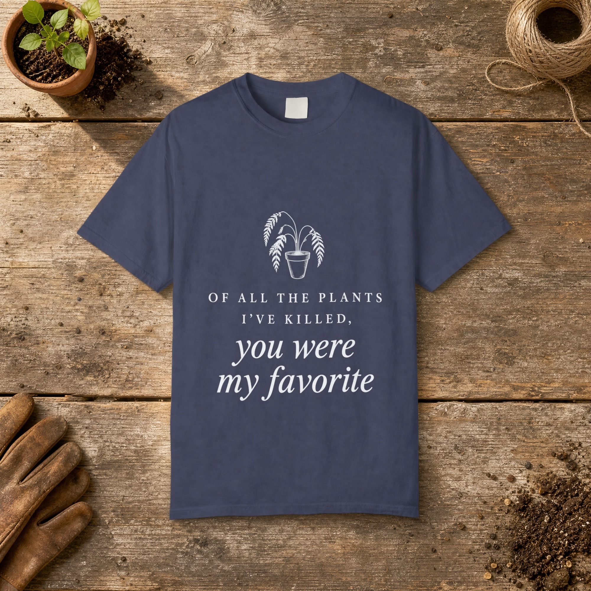 Of All the Plants I've Killed gardening t-shirt in Denim, laid flat on a potting bench with gardening gloves and a trowel
