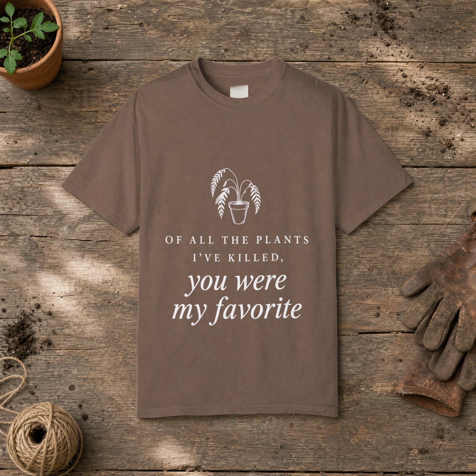 Of All the Plants I've Killed gardening t-shirt in Espresso, laid flat on a potting bench with gardening gloves and a trowel