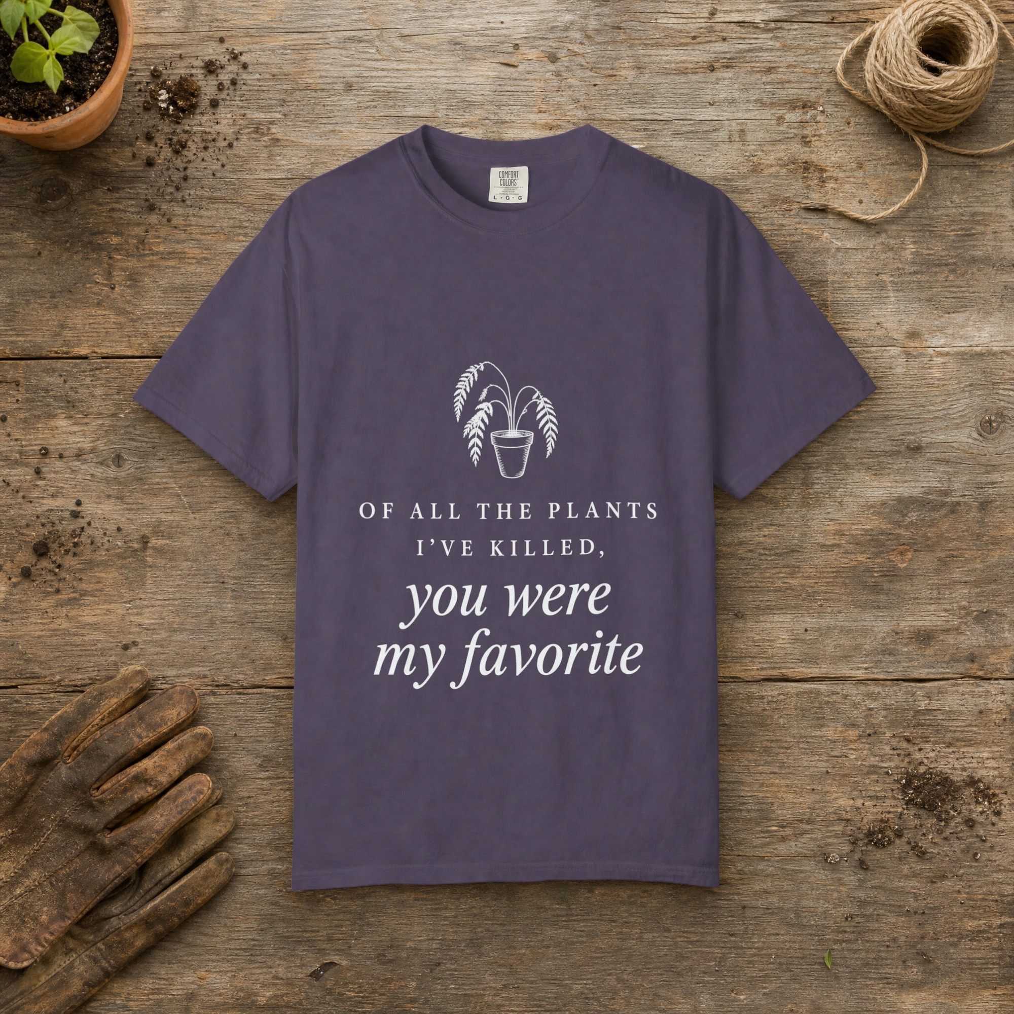Of All the Plants I've Killed gardening t-shirt in Wine, laid flat on a potting bench with gardening gloves and a trowel