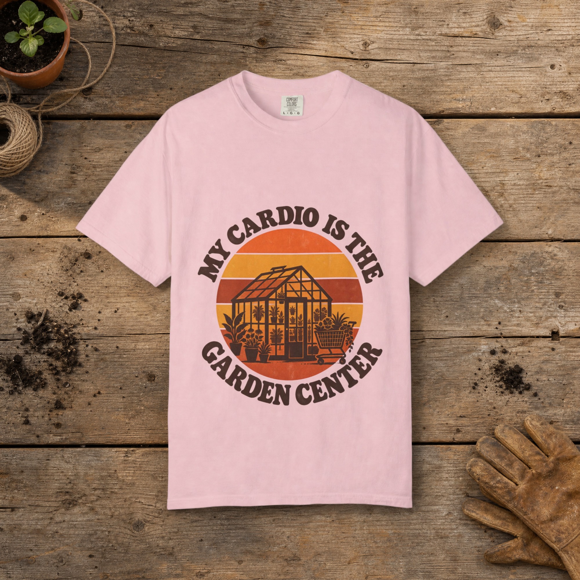 My Cardio Is the Garden Center gardening t-shirt in Blossom, laid flat on a potting bench with gardening gloves and a trowel