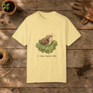 I Live Here Now gardening t-shirt in Butter, laid flat on a potting bench with gardening gloves and a trowel