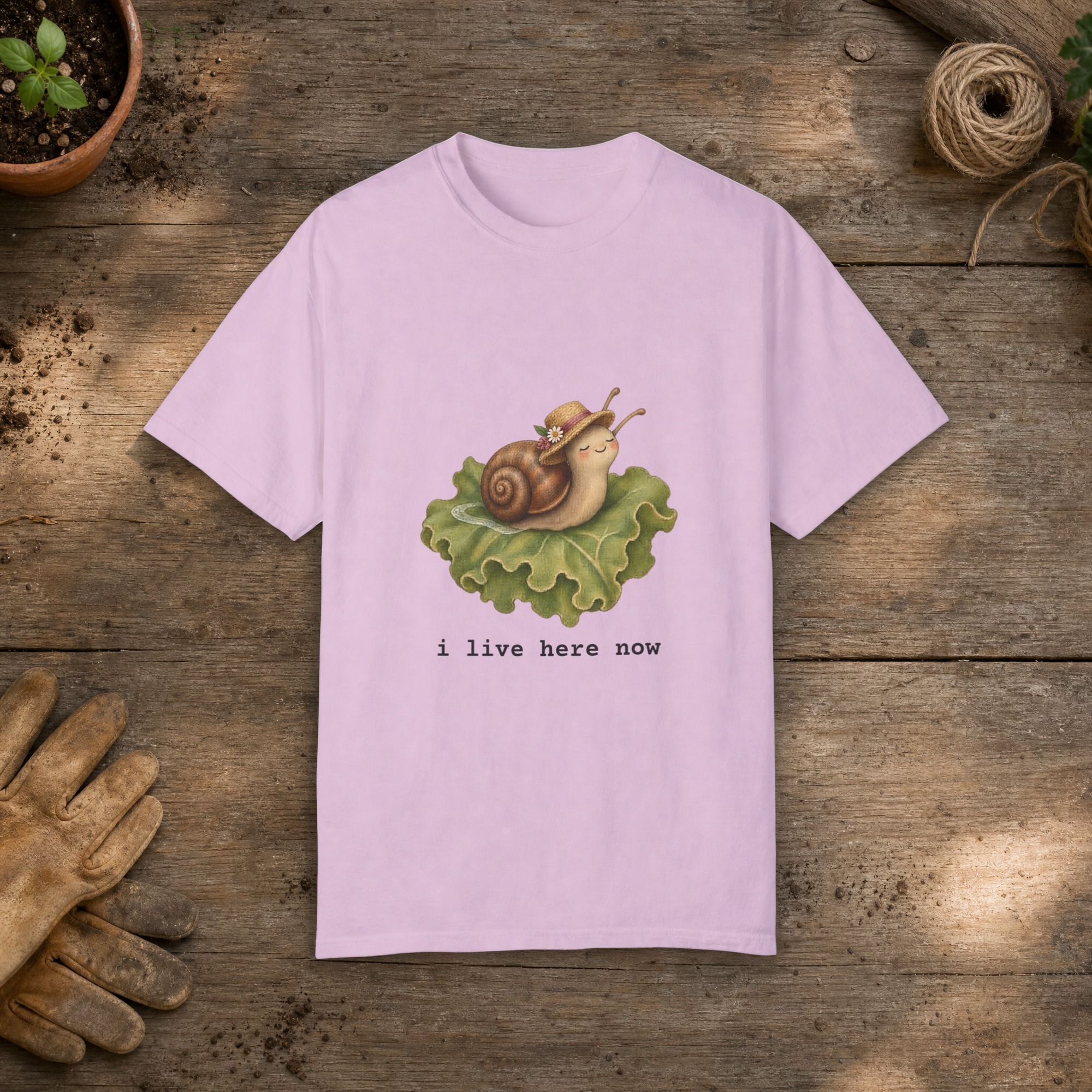 I Live Here Now gardening t-shirt in Orchid, laid flat on a potting bench with gardening gloves and a trowel
