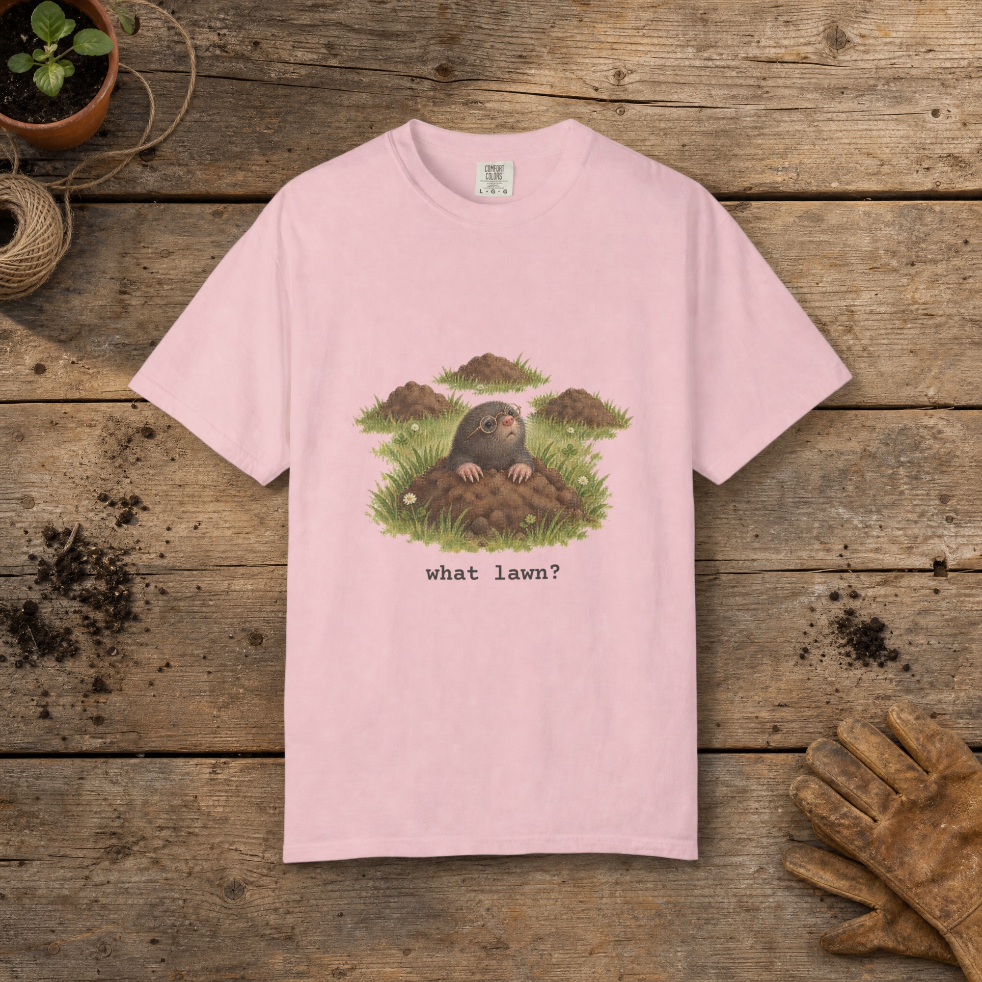 What Lawn gardening t-shirt in Blossom, laid flat on a potting bench with gardening gloves and a trowel