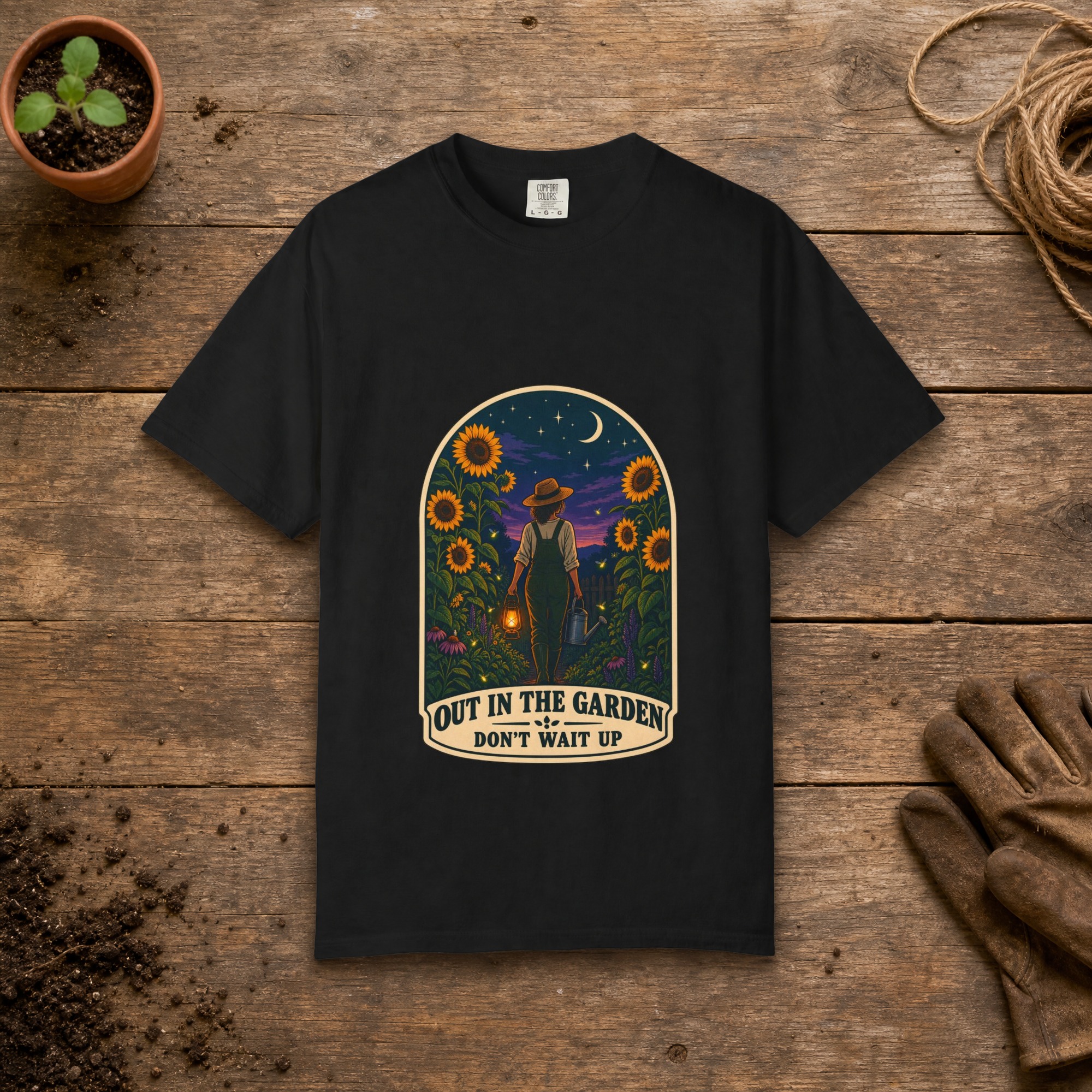 Out in the Garden gardening t-shirt in Black, laid flat on a potting bench with gardening gloves and a trowel