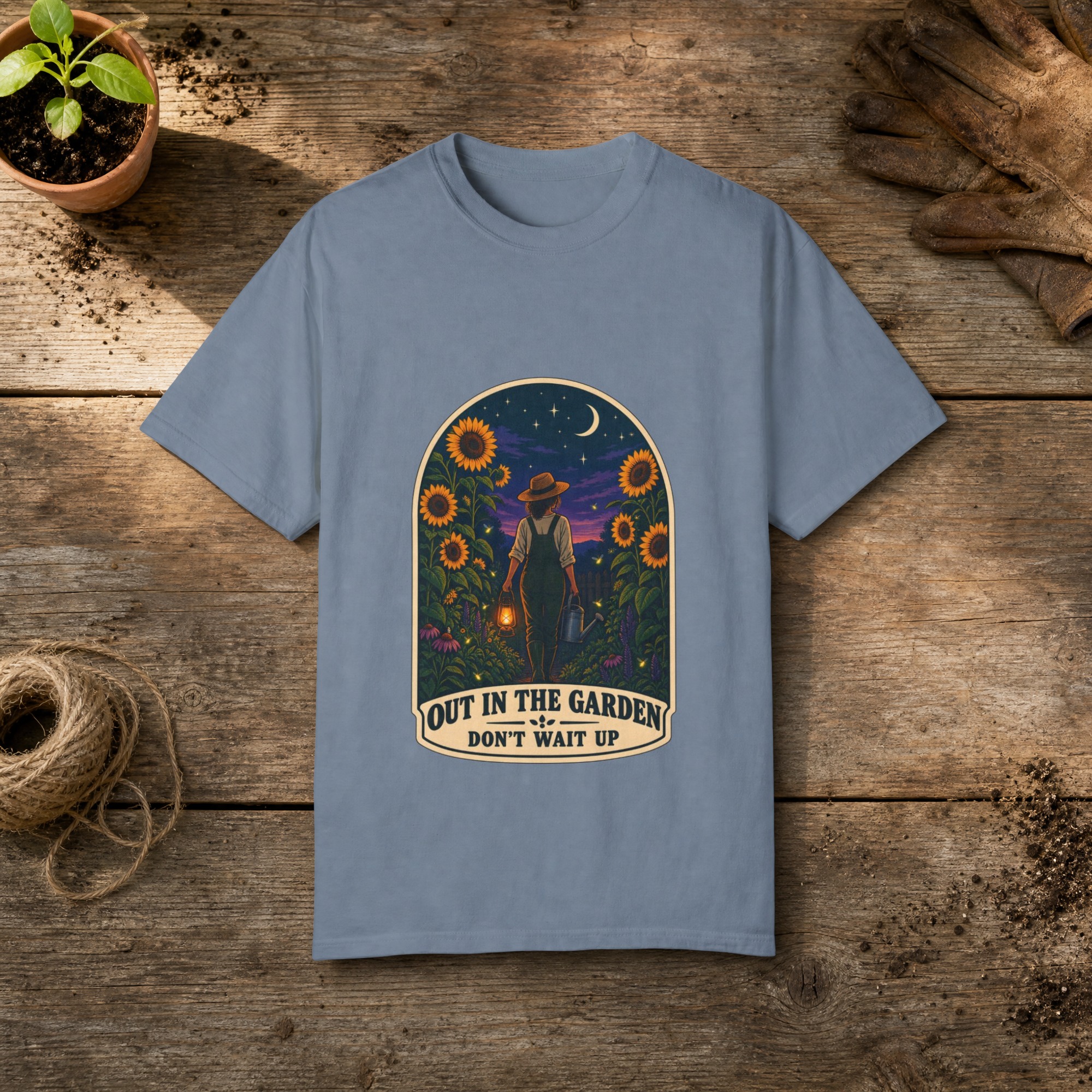 Out in the Garden gardening t-shirt in Blue Jean, laid flat on a potting bench with gardening gloves and a trowel