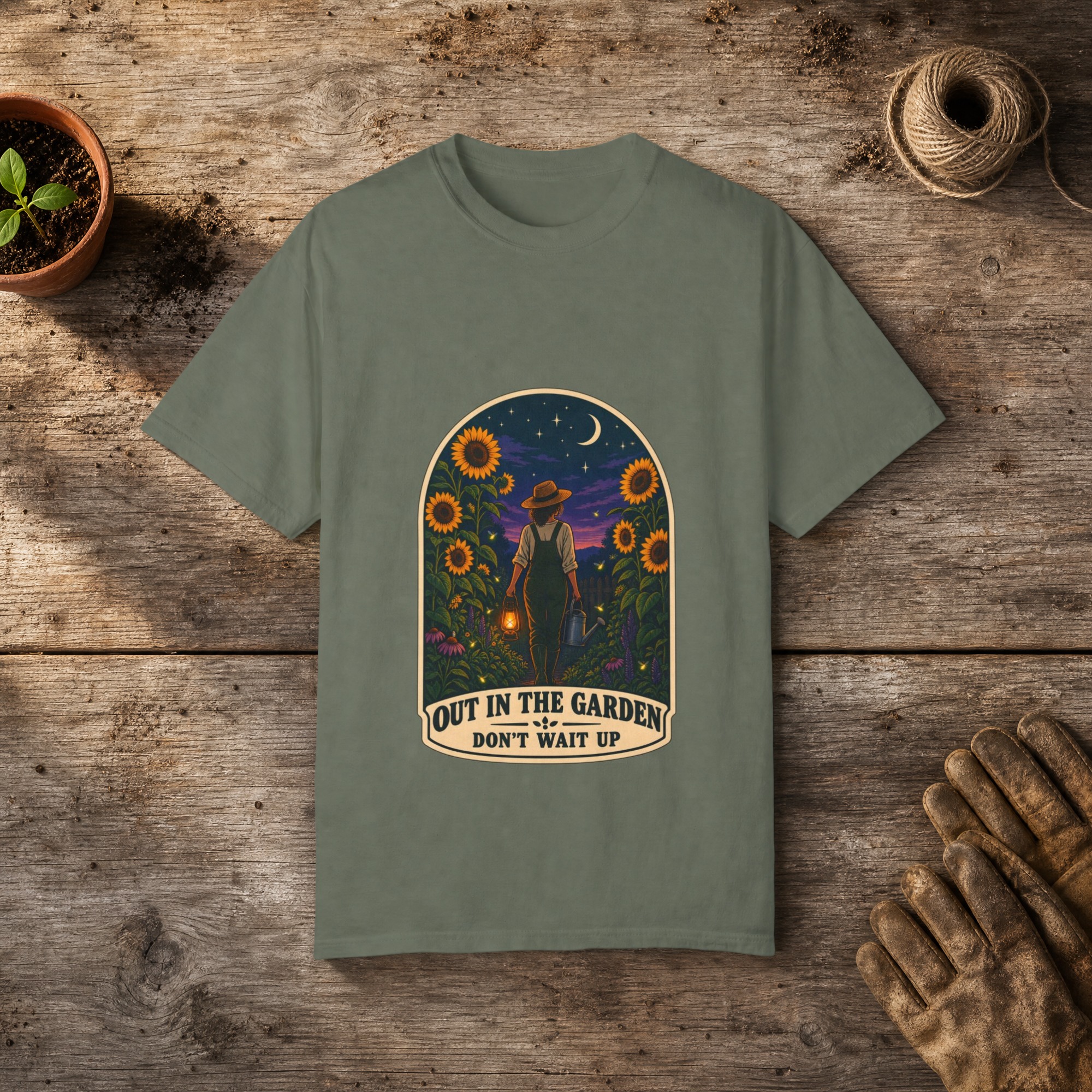 Out in the Garden gardening t-shirt in Moss, laid flat on a potting bench with gardening gloves and a trowel