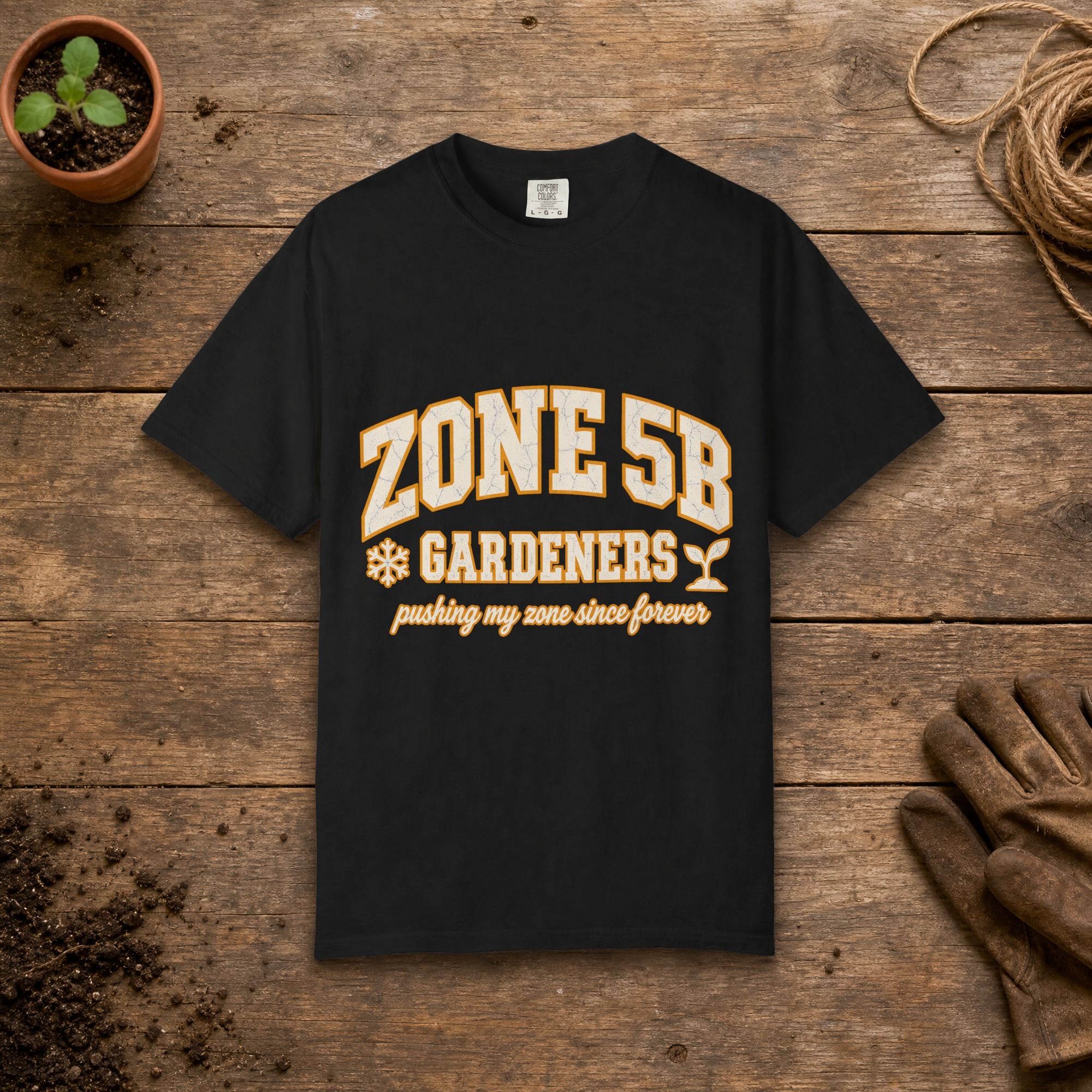 Zone 5B Gardeners gardening t-shirt in Black, laid flat on a potting bench with gardening gloves and a trowel