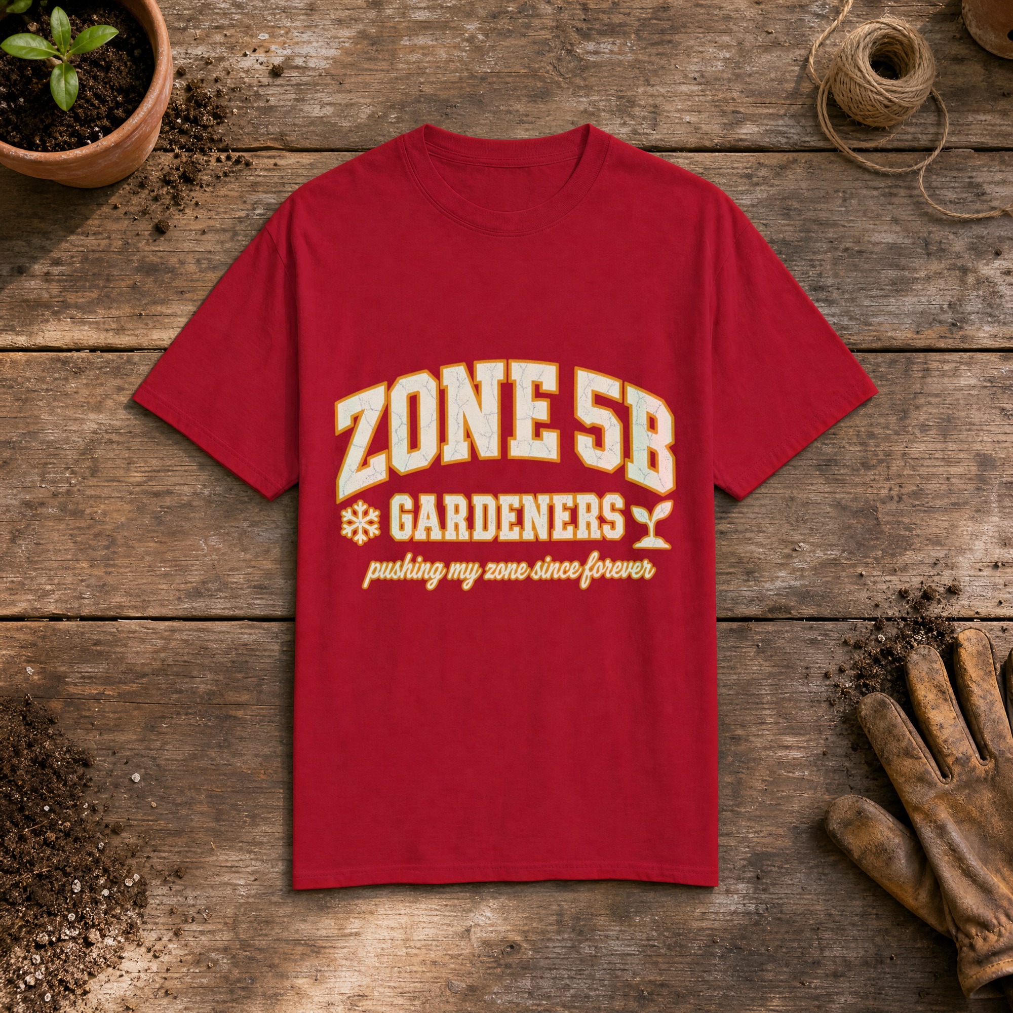 Zone 5B Gardeners gardening t-shirt in Red, laid flat on a potting bench with gardening gloves and a trowel
